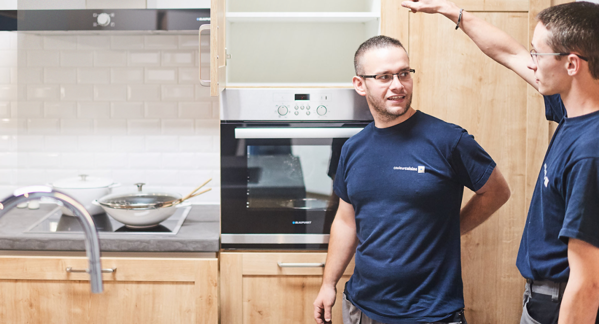 Deux installateurs de Couleur Cuisine discutent lors de l’installation d’une cuisine équipée en bois. Un four encastré est visible, ainsi qu’un plan de travail avec des ustensiles de cuisine.